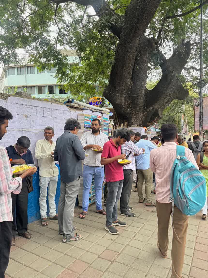 People lined up along a compound wall eating from paper plates