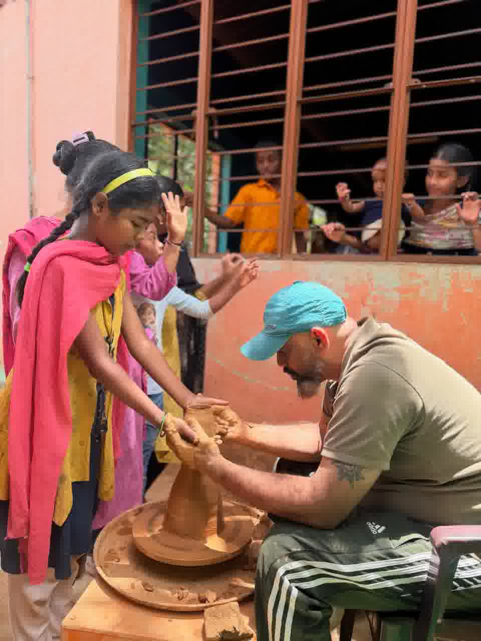 A potter at a portable wheel guiding a young girl in a yellow dress as she shapes a clay vase
