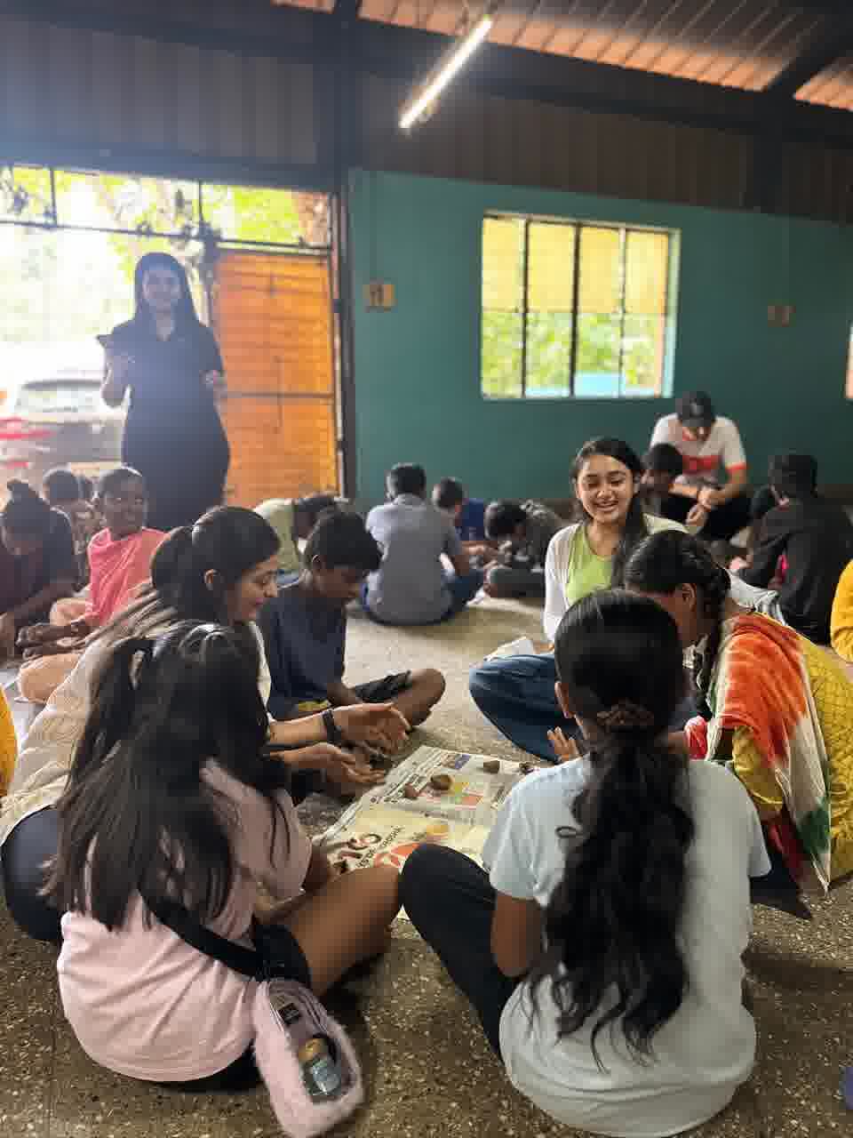 A wider view of the clay session, children seated in small groups over newspaper while an instructor watches from the doorway