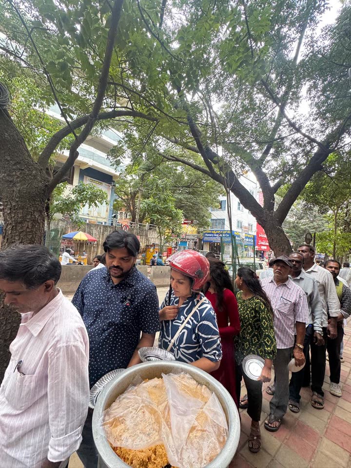 A queue of people with plates waiting under the trees for their meal